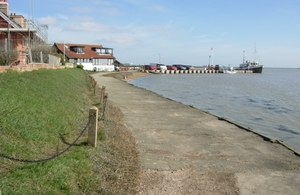 Image shows path curving from right next to the harbour past some houses towards a jetty with a boat moored at the end