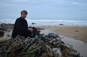 Image shows a young man with a pile of rope and fishing nets to the left with a view over the sand to the sea to the right