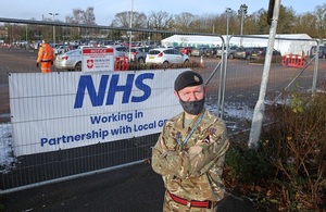 Man in British Army uniform standing with his arms crossed in front of a sign that reads NHS.