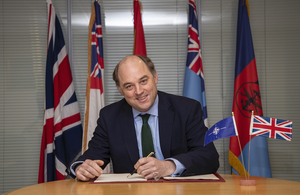 Defence Secretary Ben Wallace sitting at a desk, pen in hand, smiling at the camera in front of 4 different flags.