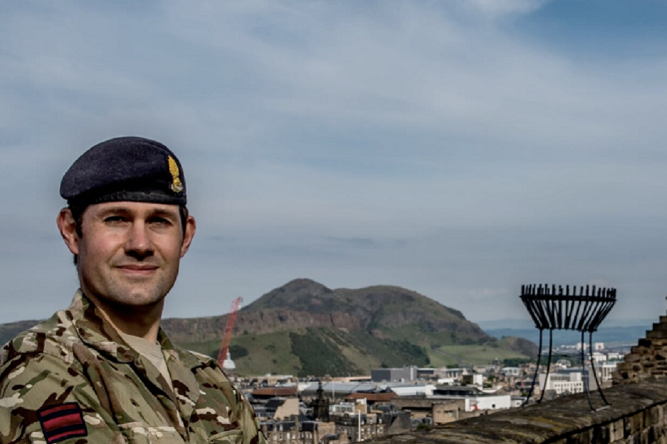 Pictured on the left hand side of the image in uniform with buildings and landscape in view behind him. 