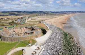 Image shows circular defensive structure to the lower left of the picture protected by a bank of rocks running in front towards a long beach stretching into the distance