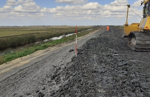 Image shows a stream running diagonally from the left across flat fenland, with a raised bank of stones running parallel under construction. A man in high visibility clothes and a digger can be seen to the far right