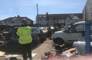 Image shows an Environment Agency officer wearing a high visibility jacket looking across yard full of vehicles being broken down for recycling, which are being inspected by British Transport Police officers. A row of houses can be seen outside the yard