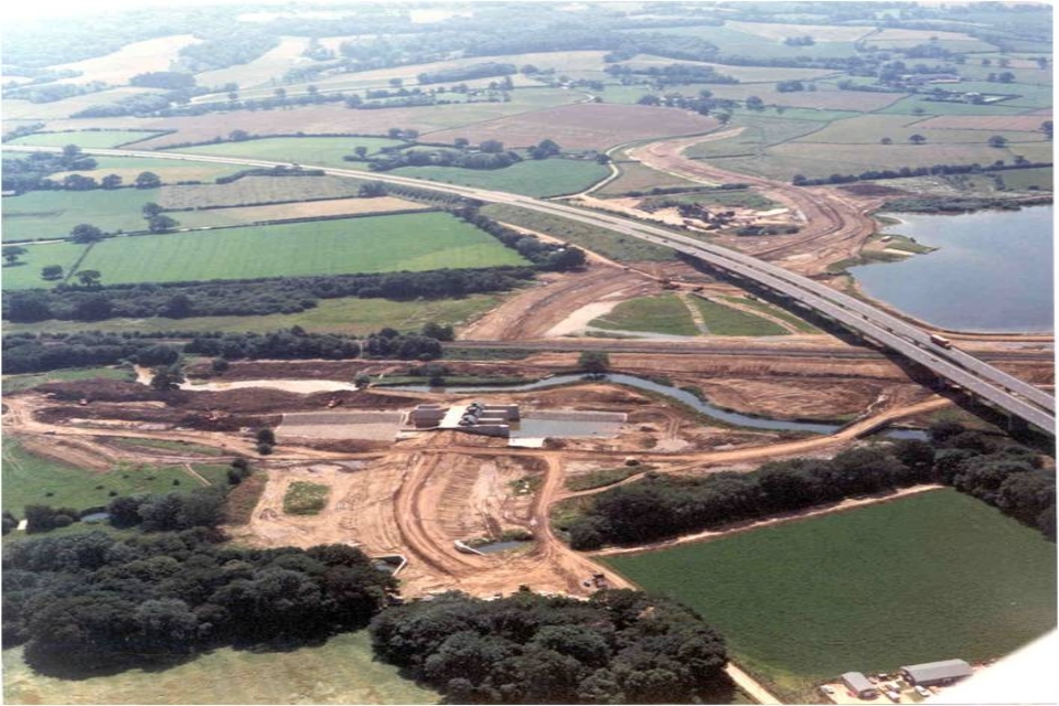 Image shows aerial view of site under construction in flat landscape of fields. A motorway runs diagonally across the right of the picture