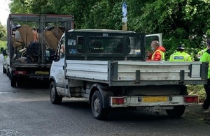 Image shows small white tipper truck to right with three people in high visibility jackets to one side, parked behind another truck with a metal cage filled with rubbish