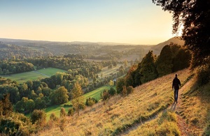Image shows a person walking to the right of a picture overlooking a landscape of trees and fields in the valley below