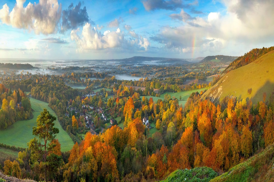 Image shows a tall hill lit by the morning sun to the left with above a landscape of autumnal trees. There is mist above the fields in the distance and a rainbow can be seen to the right