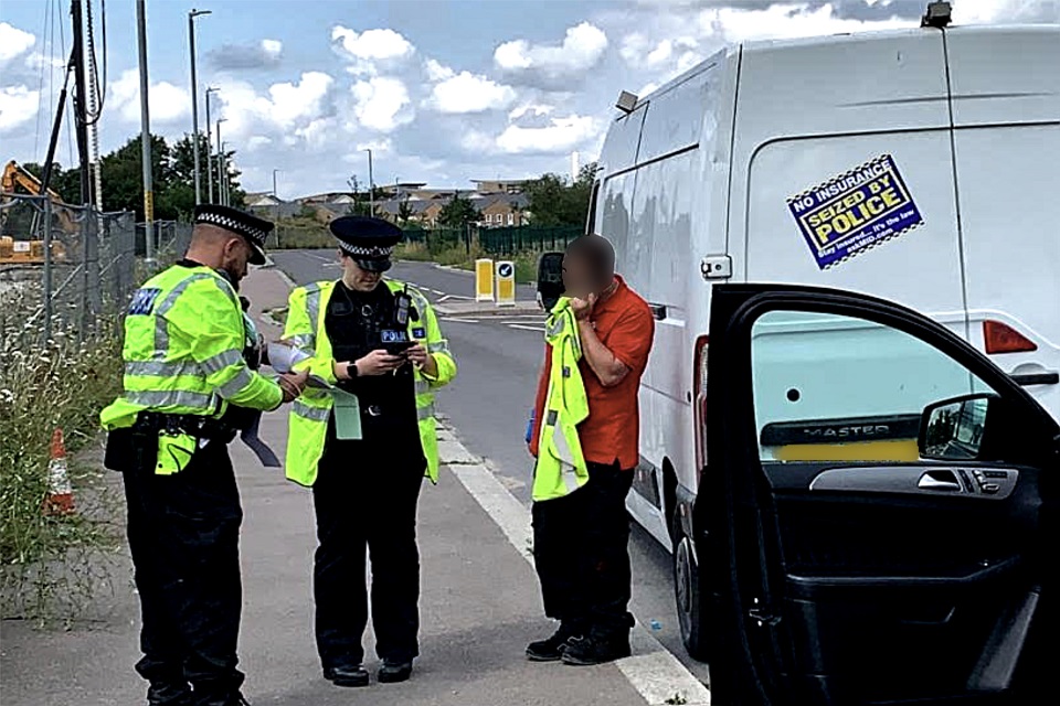 Two police officers to the left check the paperwork of a driver standing next to a white van to the right