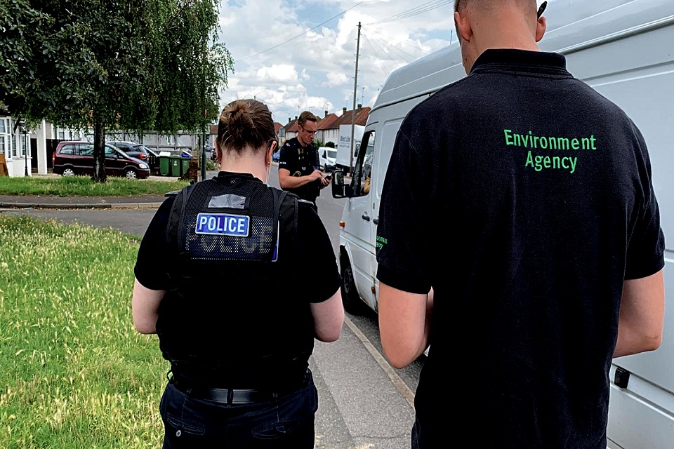 Image shows a man checking the paperwork of the driver of a white van, viewed between a police officer and an Environment Agency officer 