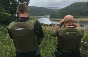 Two environment agency officers facing a riverbank with binoculars.