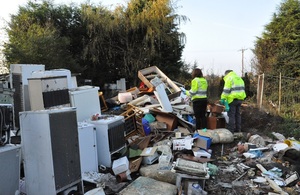 Image shows 2 environment agency officers dressed in high visibility jackets to the right of the picture, inspecting a pile of waste including old fridges and furniture
