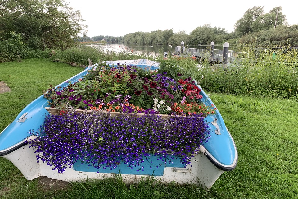 A rowing boat, pictured from behind, sits on a grassy bank near the river. The boat is filled with a colourful array of flowers, which spill over the sides of the boat. The river can be seen in the background.