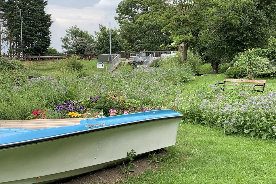 A side view of a rowing boat sitting on grass. The boat is filled with flowers. Around it is grass and more wildflowers in an open green space with a bench.