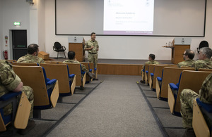 Maj Gen Roe, Commandant of the Defence Academy, standing in the lecture theatre talking to seated MPs and Peers dressed in Service uniform, with a screen behind showing the text 'Welcome Address'.