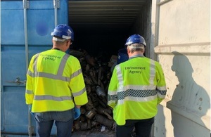 Image shows two Environment Agency officers wearing high-viz jackets and hardhats looking through the door of a shipping container half full of catalytic converters