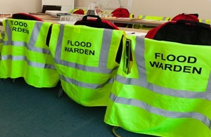 На изображении показаны три хорошо заметных куртки с надписью «Стражи наводнений», висящие на спинках стульев в зале заседаний.
