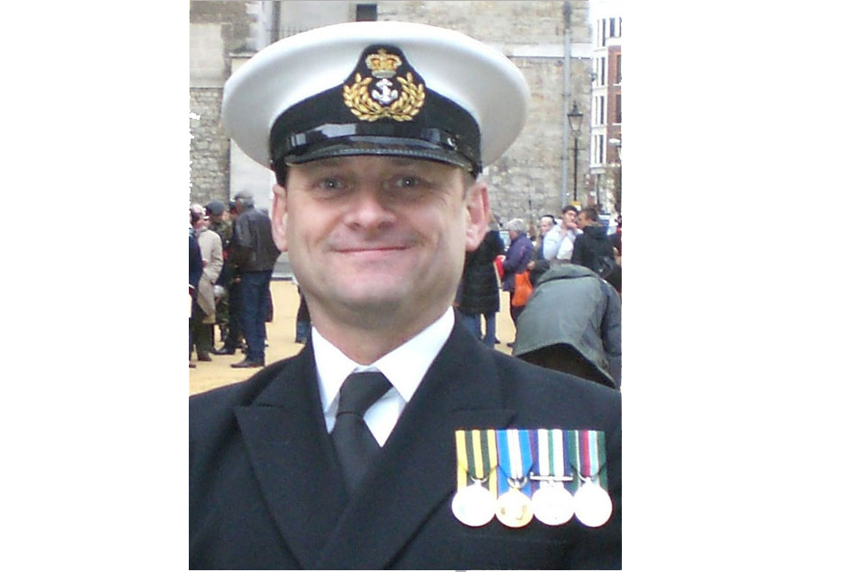 Tony Matthews, dressed in Royal Naval Reserve uniform and decorated with 4 medals, smiles into camera.