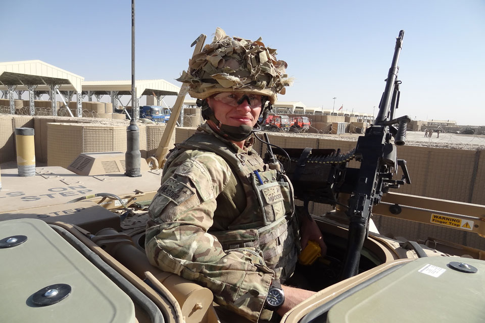 Colin Murray dressed in khaki camouflage, pokes out the top of a tank and holds a machine gun. He is smiling into camera.