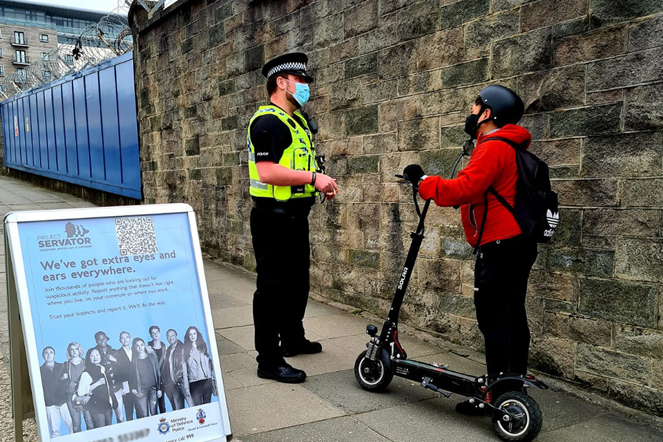 Project Servator officer, wearing a high vis jacket and face mask, talking to a person on a scooter wearing a red jacket, with a stone brick wall and fence line in the background and a Project Servator poster in the foreground.