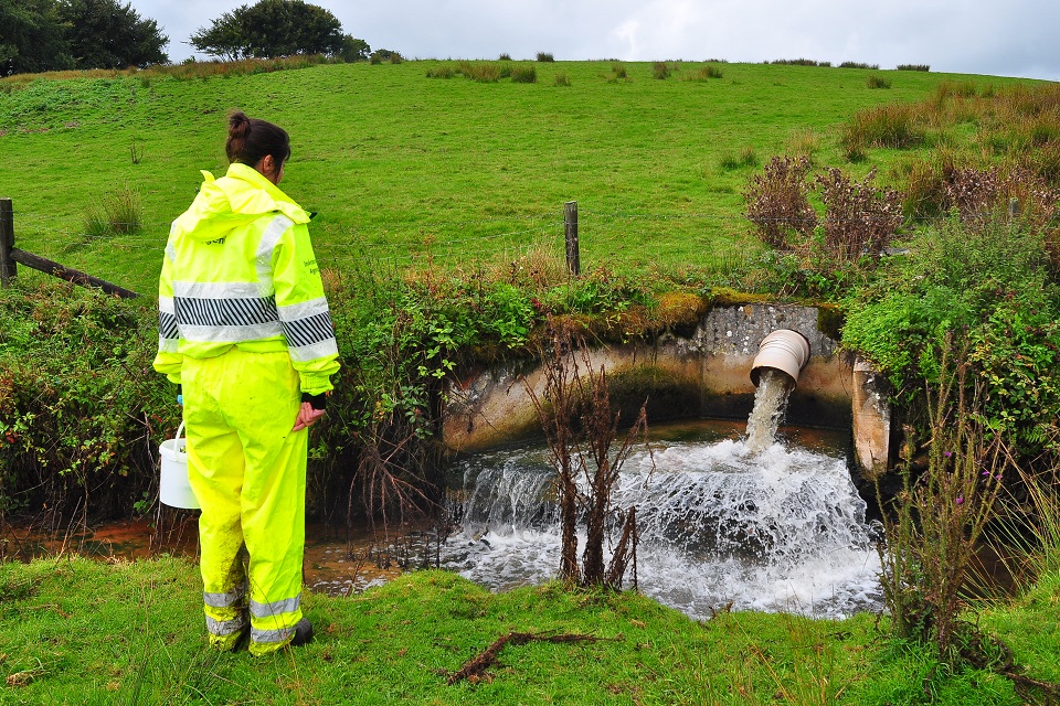 A female Environment Agency officer is next to a pipe discharging into a small river