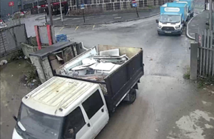 На изображении показан черно-белый самосвал, полный отходов, проезжающий через металлические ворота. Два других грузовика можно увидеть припаркованными на дороге сзади.