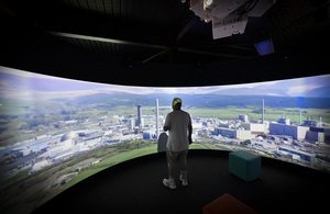 Мужчина в белой футболке и джинсах стоял перед большим проекционным экраном, показывающим аэрофотоснимок участка Селлафилд.