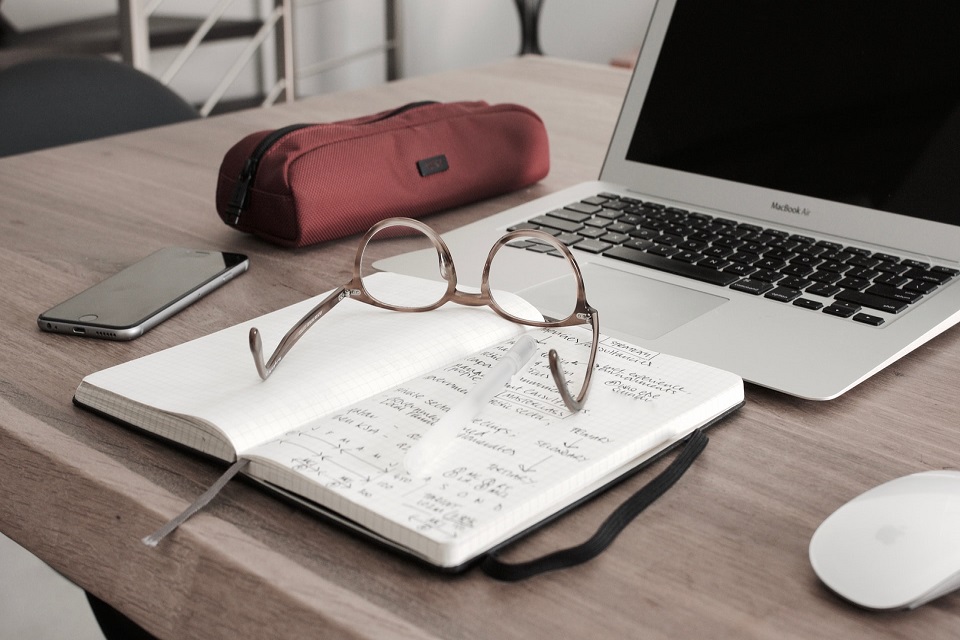 In the foreground, spectacles on top of an open notebook in which there is writing in black ink. Behind these items there is an open laptop.