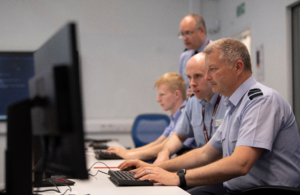 A side-on image of a man sat at a keyboard with people next to him out of focus