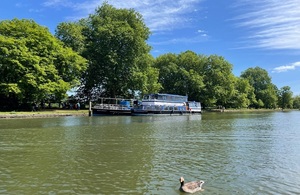 Image shows two river cruisers moored at a riverbank next to some trees. A duck can be seen in the foreground.