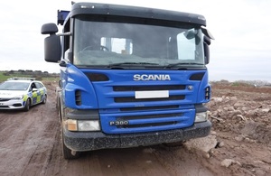 Image shows front view of blue truck parked on muddy ground with police car to the left.