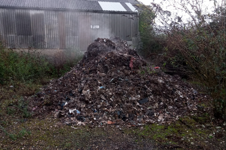 Image shows a large pile of waste with smoke rising from its top in front of a warehouse.