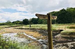 mage shows a person walking from left to right through countryside following a path beside a dried-up stream towards a small footbridge. The foreground is dominated by a sign pointing left and right to 'England Coast Path'I