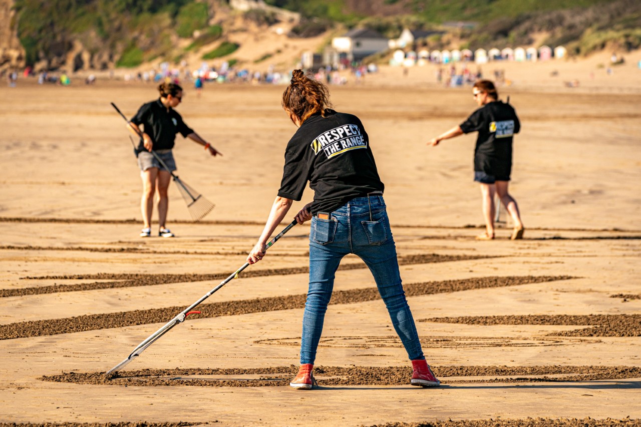 Three sand artists wearing t-shirts saying 'Respect the Range' rake sand on the beach at Saunton Sands