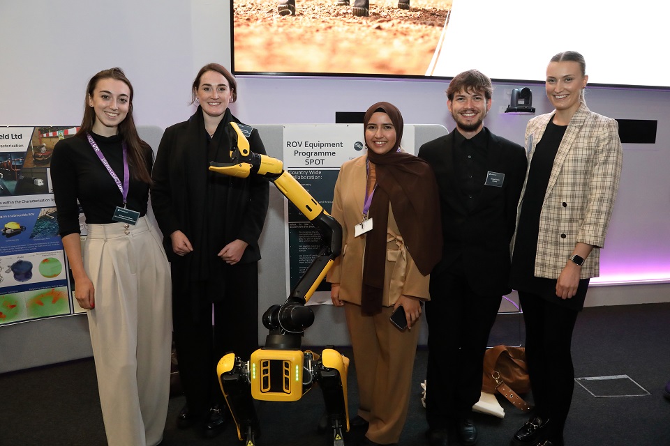 Five graduates stand in a line with a robotic dog