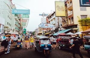 Busy street scene in Thailand. A tuk-tuk is in the centre of the street and people are walking in the road as well. The street is lined with shops with each one having large signs outside.