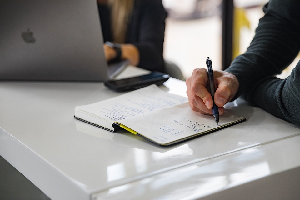 Man writing in notebook. Blurred out image of a woman near him, working on a computer.