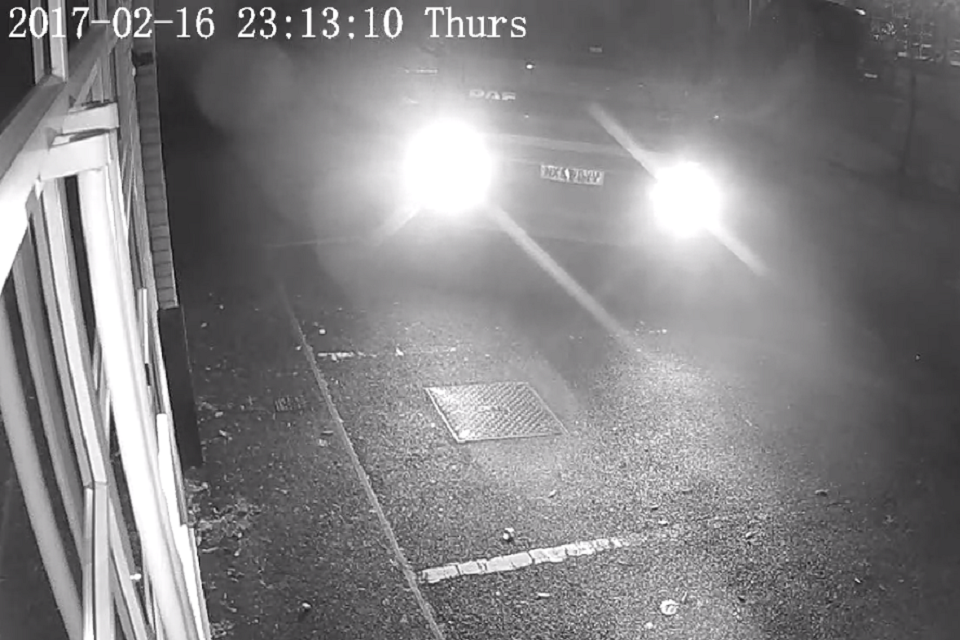 A black and white CCTV image shows the headlights of a truck coming into view 