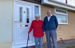 A woman and a man stand by a front door. The woman has white hair and is resting on a walking stick. The man is slightly taller than the woman/.
