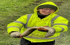 A man wearing glasses and dressed in a yellow high-visibility jacket holds a large eel to the camera