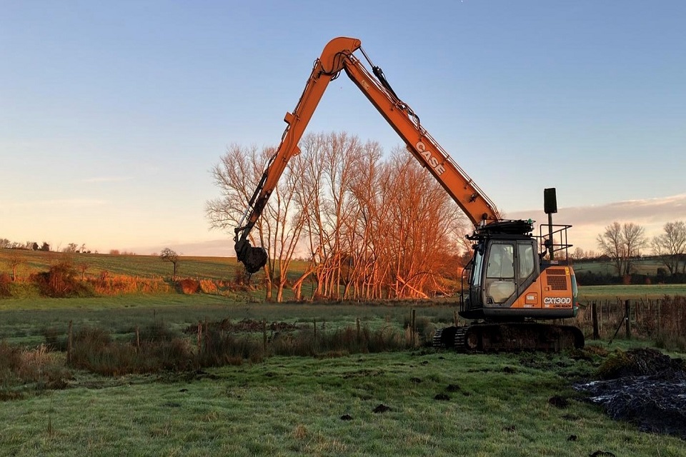 An orange and black excavator can be seen leaning over a river, with a tree is the distance, under blue and reddy skies