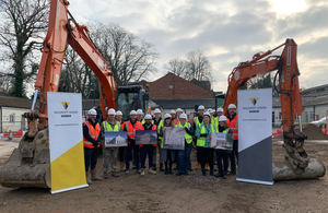 A group of 17 people in high vis stand between two orange diggers on an area of open earth. Four of them hold printed artist’s impressions of the new building. The group are flanked by two Wilmott Dixon banners.