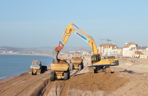 A sandy-coloured excavator puts shingle into a lorry of the same colour. Another lorry and a bulldozer work alongside. Calm seas to the left below sunny skies. Houses and flats are in the middle and long distance