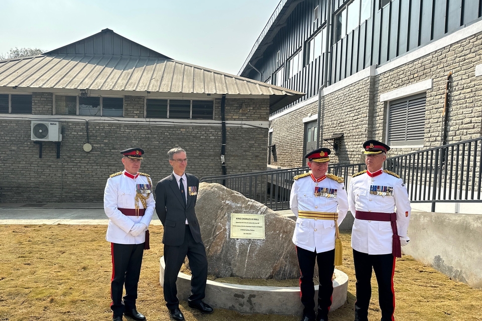 Three men in ceremonial military attire and another wearing a suit, stood in front of a stone plaque with an inscription headed ‘King Charles III Hall.’