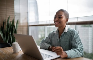 Smiling woman sitting at a table and in front of a laptop. To her right is a mug and a plant.