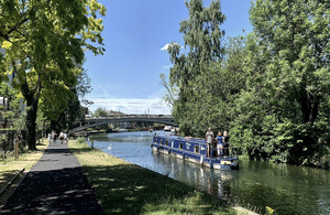 A blue narrowboat is seen on a river, surround by trees in full leaf and sunny skies