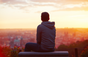 An image of a man looking out at a town view is pictured. It is sunset. He wears a hoodie
