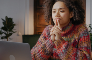 A woman is sat in front of her laptop. Her expression is contemplative.
