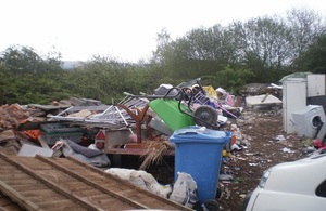Assorted waste and rubbish, such as an old washing machine, soil, a fence panel, a wheelbarrow, metal railings and bricks are between the camera and a hedge and grey skiesa