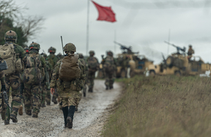 Image of dozens of soldiers marching towards a red flag and two vehicles with mounted weapons.